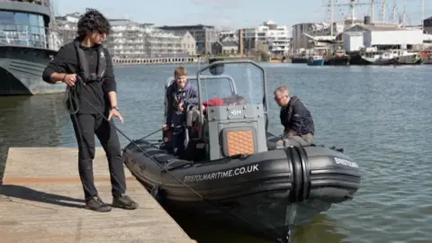 A young man holds the rope of a RIB speed boat alongside a wooden pontoon. 