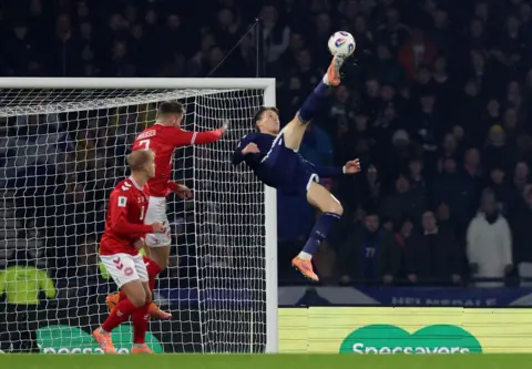 Reuters Scott McTominay kicks the football in an overhead bicycle kick. He wears the navy Scotland football strip. Two Denmark players stand behind in him in red football strips. The net can be seen behind him as well as many fans staring at Scott.