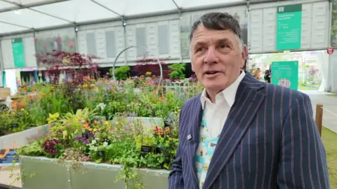 A wide shot of Nick Wade standing in front of a flower display. He is wearing pin-striped navy jacket and a flower waistcoat. He has short dark hair.
