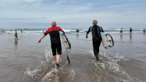 Two men in wetsuits are walking into the sea at Bude, Cornwall. they are both holding surf boards and 