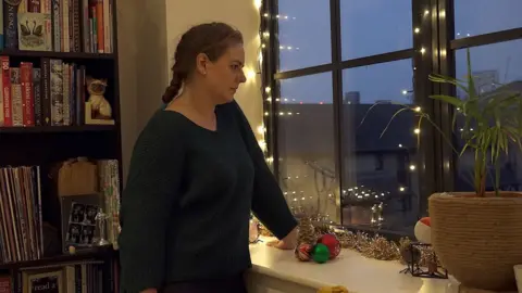 Kate Howe stands in her flat, looking out of the window. She is wearing a v-neck dark jumper and her hair is in a plait . There are fairy lights around the window.