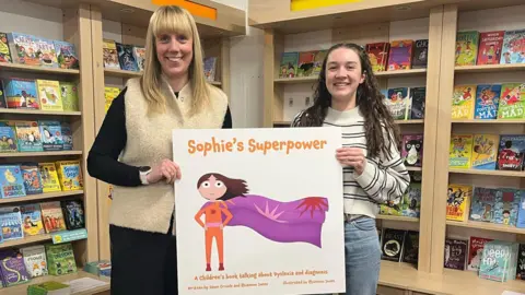 Dawn Crowle and Rhiannon Jones. Dawn has long blonde hair and is wearing black with a cream knitted vest. Rhiannon has long dark hair and is wearing blue jeans and a striped jumper. They are stood side by side, holding a large copy of the Sophie's Superpower book cover. It is white with an illustrated superhero wearing an orange suit with a purple cape. 