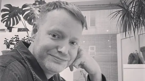 Chris with his head resting on his hand smiling at the camera with a plant in the background