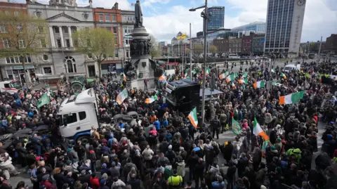 PA Media A crowd, several of them waving Irish Tricolour flags stand in O;Connell Street in Dublin. Two lorries are visible in the midst of the crowd