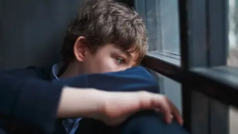 A file photo of a dark-haired boy sitting on the floor by a grey-framed window, wearing a light blue shirt and dark blue sweater. His arms are folded and balanced on his knees. He is looking towards the window with a haunted expression.