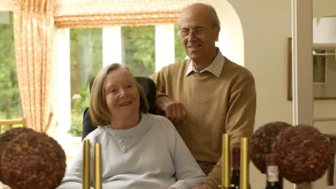 Margaret Tebbit sitting down, while her husband Norman stands next to her. 