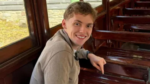 Alfie is sitting inside a traditional Manx electric tram, on oak wooden benches, with windows along the left. He is smiling, has short slightly wavy brown hair, and is wearing a light brown sweatshirt.