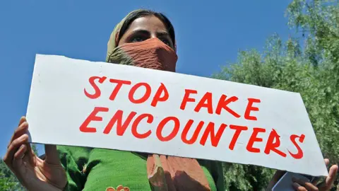 Getty Images A woman wearing a green tunic protests against "encounter" killings in Indian-administered Kashmir. She's holding a board saying 'stop fake encounters' and half her face is covered by a brown-and-green shawl 