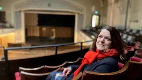 BBC Lynn is sitting in a theatre chair looking back over her shoulder at the camera. She has brown shoulder length hair. She is smiling and wearing a red scarf. The stage is blurred and in the background.