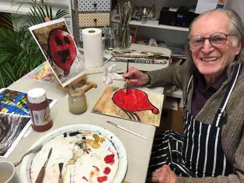 A man with greying hair tied back sits at a table with a painting of a red balloon on front of him. He is holding a paintbrush and smiling at the camera.