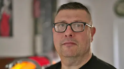 Brian Buckle looks straight towards the camera - he is wearing square glasses and a black jumper. He has short dark hair and dark stubble, and is pictured in a kitchen.