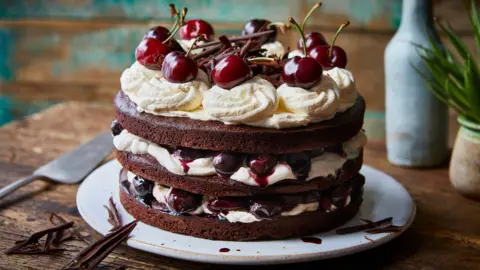A Black Forest gâteau cake - with three layers of chocolate cake, cream and cherries - sits on a white plate on a wooden table 