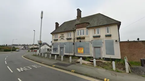 The boarded up pub stands on a corner of a road. It is beige in colour with metal covers on lower ground windows. There are small bollards outside it on the bend in the road.