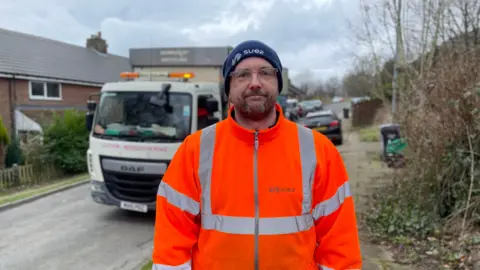 Mark Ansell/BBC Bin man Jack Ford in orange high visibility jacket in front of a bin lorry with a blue hat and glasses.