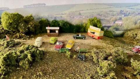 Gerry Lodge A close up of a model hillside with vintage cars parked along with a small wooden stall and ice cream van. There are shrubs and trees and more rolling hills seen in the background