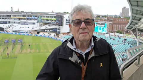 Lily May-Symonds/BBC Graham Gouldman, who has grey hair and is wearing glasses and a blue raincoat, stands on a balcony at The Oval. The pitch and the stands can be seen behind him.