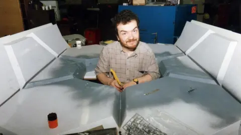 Mike Tucker A younger Mike Tucker, with dark hair and beard, is smiling from the middle of a large spaceship model he is building.