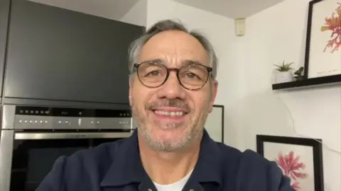 Guy Whittingham smiling at the camera. He has grey hair, green eyes and dark framed glasses. He is wearing a navy jumper with a collar and a white t-shirt underneath. In the background are grey cupboards and pink flower pictures.