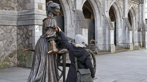 peterspiro/ Getty Images A woman touching the breast of the Molly Malone statue in Dublin.   The woman has her back to the camera and is wearing a dark overcoat, a grey woolen hat and a grey shoulder bag. 