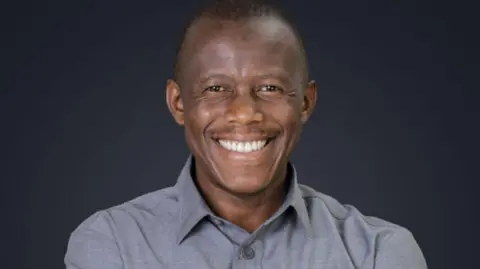 The Citizen Humphrey Polepole smiles. He is wearing a grey shirt.