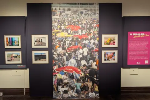 Jadzia Samuel/BBC Some of the art provided for JJ Waller's Beside the Sea exhibition at the Hastings Museum and Art Gallery. A huge vertical picture of people on the beach hangs in the middle of the image, with a number of framed pictures seen either side. A pink display outlining the exhibit can be seen on the right-hand side of the image. All pictures are set against a purple wall.
