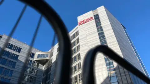 Emma Baugh/BBC Image of outside of the hotel, there is a red sign with Hilton Garden Inn, the hotel is behind black barriers.