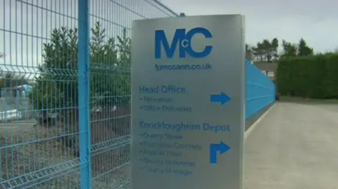 FP McCann sign. It is grey with blue writing, beside a blue fence.