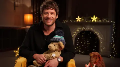 James Norton - who is wearing a black t-shirt and beige chinos - is smiling, while holding a teddy bear. The teddy bear is wearing a small glucose monitor. There is also a cuddly toy mammoth beside him.
