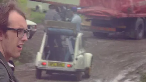 Mensch Films/Robin Mahoney A vintage image of the camera crew in an open-topped car standing up through the roof with a large camera. Robin is in the foreground, his face visible on the left. He is looking to the right, off camera. The ground is very muddy and there is a lorry with a red tarpaulin in the background.