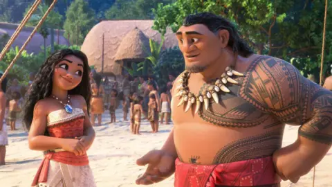 A still from Moana 2, with the main characters on a Polynesian island, with lots of different characters on the beach in the background. There is a straw hut and lots of trees.