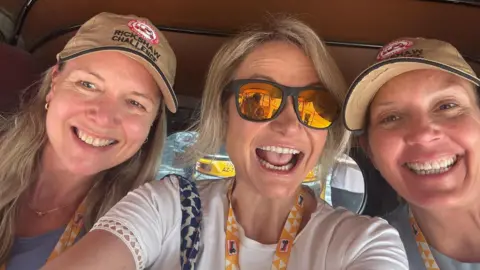 Claire Jenkins Three middle aged women with blonde hair in the back of a tuk-tuk. Two are wearing caps and another is wearing black-framed sunglasses with reflective orange lenses. All of them are smiling.