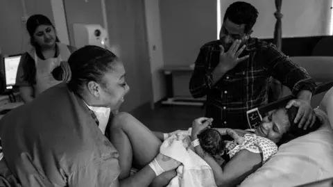 James Clifford Kent A black and white photo of a new mother lying in a hospital bed holding her newborn, looking up at her partner who is visibly emotional, while a smiling midwife looks on.