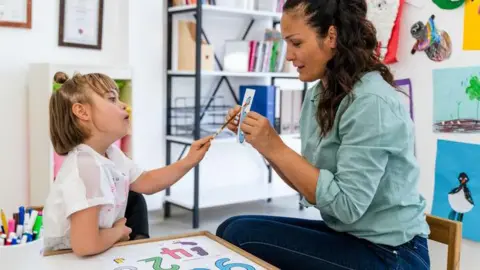 A pupil and a teacher are in a classroom doing maths. The teacher who has long dark hair is holding up a piece of paper and the girl, who has a white top and her hair tied back, is pointing at it. There are books on shelves behind them, with notices in frames and artwork on the walls.