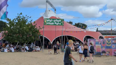 People walking by the Feel Good Food Hall tent at Truck Festival. There is a group sitting beside it. There is a colourful mural on the right. It is cloudy.