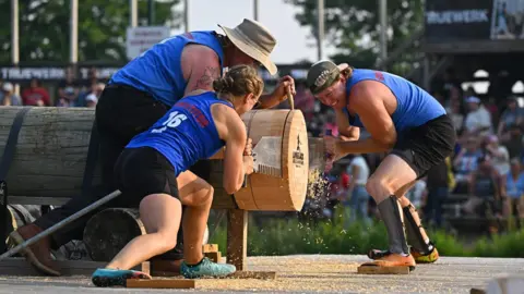 Lumberjack World Championships Two women brace themselves and crouch on either side of a huge wooden log while using a double-handled saw to slice through it