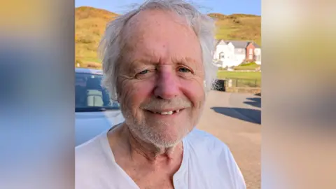 David Patterson David Patterson is standing on a road in the sun with green hills behind him. He is smiling at the camera.