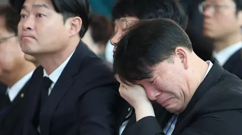 News1 A man in black suit cleans the tears in his eyes while crying. Another man by his side frowns with tears in his eyes