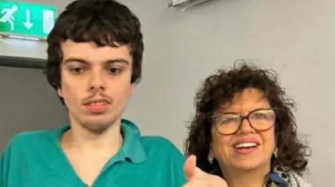 Sheenah McCann with her son Joe. Joe has short dark hair and a moustache. He is wearing a teal coloured polo-shirt. Sheenagh has curly dark hair and is wearing red framed glasses and a blue top. 