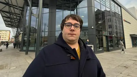 Wearside Lib Dems leader Paul Edgeworth stands outside Sunderland train station. He is wearing glasses and a duffle coat. 