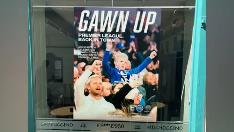 Ipswich Central A poster inside a cafe window. The poster shows a crowd of football supporters celebrating and it is focused on a young boy with his arms raised and who is wearing a blue football shirt. The words Gawn Up have been printed at the top of the photo along with additional details about an exhibition. 