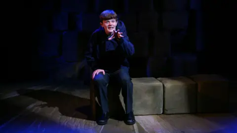 Hull Truck Theatre A boy sits alone on a low, block‑shaped bench on a dimly lit stage. Deep blue and purple lighting washes across the floor, creating dramatic shadows. Behind his is a dark, textured backdrop made of large, stone‑like blocks, giving the impression of a cellar or a cave. The boy is dressed in dark clothing and leans slightly forward, raising one hand in a poised, expressive gesture, as if performing a moment of intense focus or emotion.