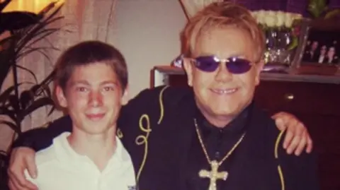 Richard Reeves Reeves with his arms around Elton John aged 14. The Elton is wearing a black outfit and a cross. They are stood near a bar.