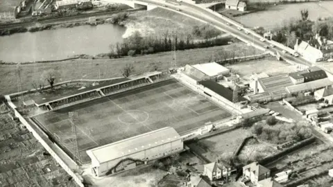 Cambridge United A black and white aerial photo of Cambridge United's Abbey Stadium between the 1930s to possibly 1950 showing the ground with two stands, four tall light towers in each corner, allotments to its left and above it a park and Newmarket Road 