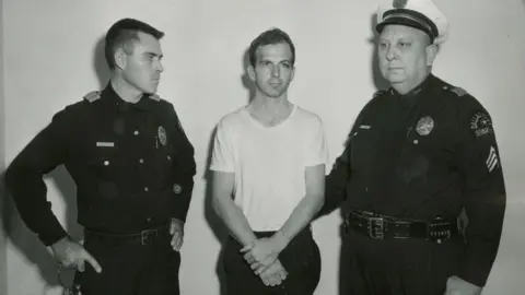 Reuters/Dallas Police Department Oswald in a white t-shirt flanked by two police officers in a black-and-white photo