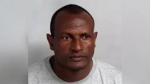 Essex Police A custody mugshot of Hadush Kebatu, who is wearing a grey sweater and has cropped black hair.