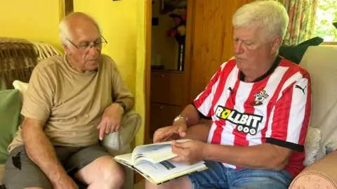 Two men sitting in chairs in what appears to be a hotel room, flicking through a book. One of them is wearing glasses, the other has a football shirt on.