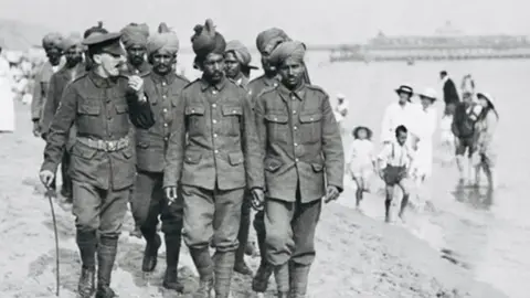 In a black and white photo from 1915, a dozen or so Indian soldiers walk on Bournemouth beach, accompanied by a British officer. The pier is in the background and people are paddling in the sea.