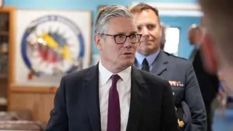 Sir Keir Starmer stood in a black suit and purple tie in an RAF base in front of a military official in the background, and the rest of the room out of focus.
