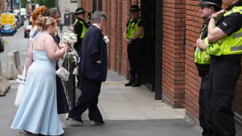 Getty Images A bride and bridesmaids arive for a wedding at the register office adjacent to and in the same building as No 10 North, Heron House as Police officers stand outside on July 24, 2026 in Manchester, England.