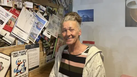 Amanda Wilkinson has greying brown hair in a bun and wears a stripy top of different shades of brown with a light hoodie.